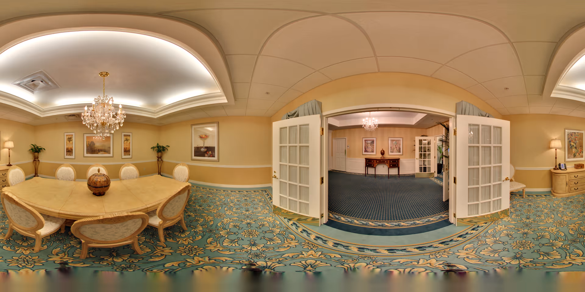 A spacious, elegant dining room with a large wooden table surrounded by eight upholstered chairs. The room features a decorative chandelier hanging from a recessed ceiling with soft lighting. The walls are painted a warm beige color and adorned with framed artwork and two potted plants. The floor is covered with a patterned carpet in shades of blue and gold. French doors open into an adjacent room with similar decor and carpeting.