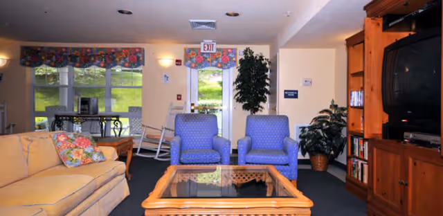 A cozy living room area with a beige sofa adorned with floral pillows on the left, two blue armchairs with a small glass-top wooden coffee table in the center, and a large wooden entertainment center with a television on the right. The room has large windows with floral valances, a rocking chair near the exit door, and several potted plants adding greenery to the space.