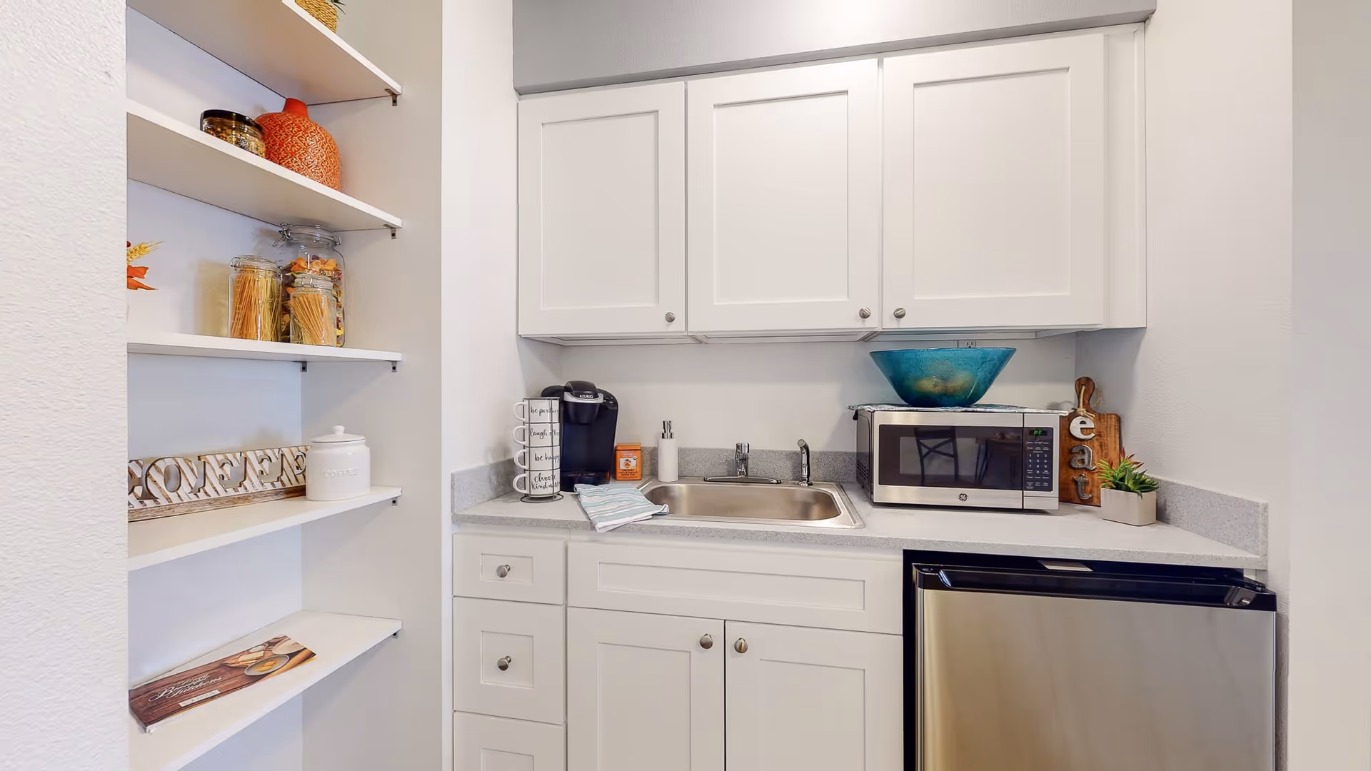 A small kitchen area with white cabinets, a stainless steel sink, a microwave, a mini refrigerator, and a coffee maker. Open shelves on the left hold jars of pasta, a decorative orange vase, and other kitchen items. The countertop has a blue decorative bowl, a soap dispenser, and a small plant.