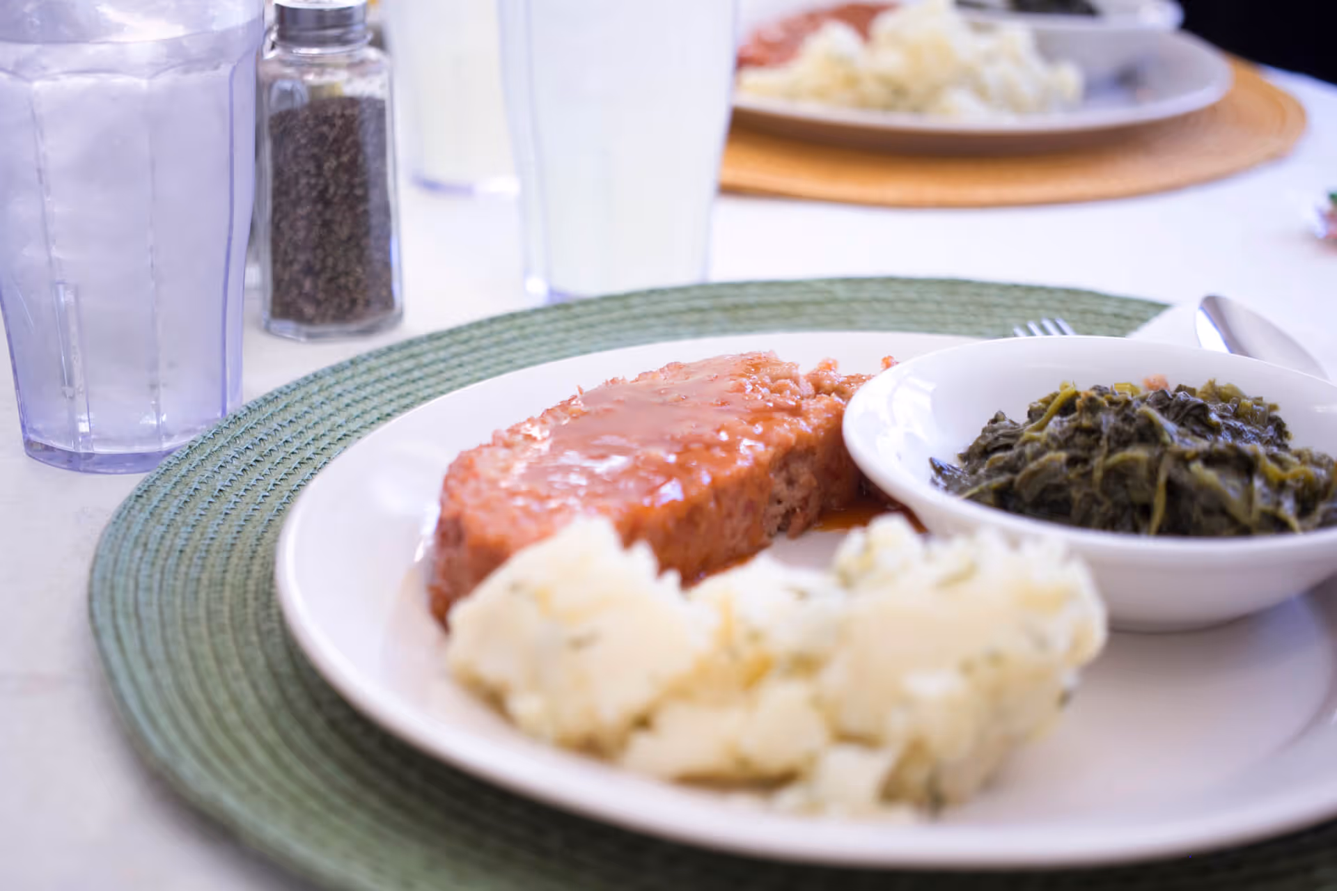 Plate of meatloaf with gravy, mashed potatoes, and a bowl of cooked greens on a table with glasses and a pepper shaker.