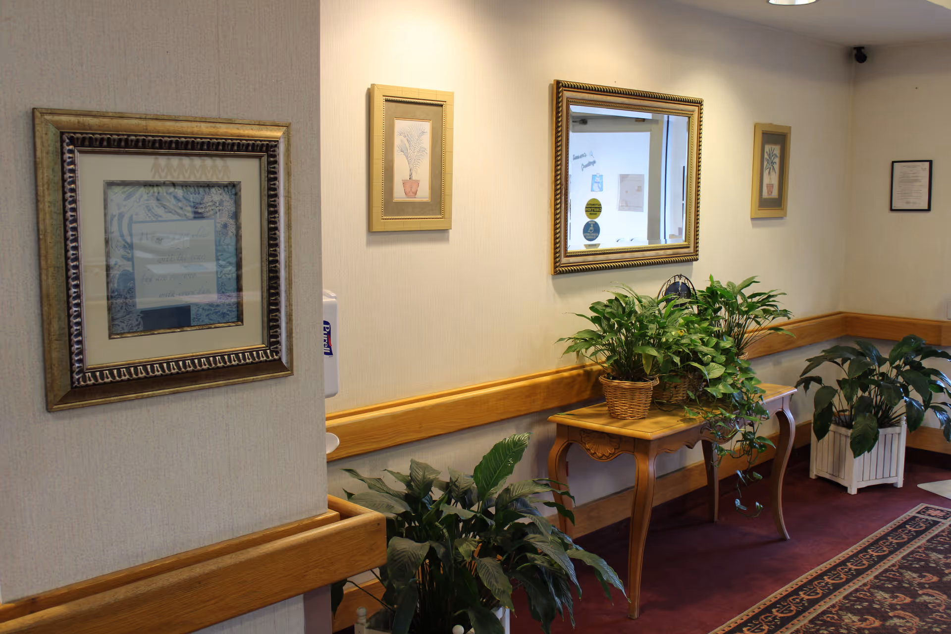 Interior hallway of a care facility with framed art and a mirror above a wooden table holding potted plants and a wall handrail.