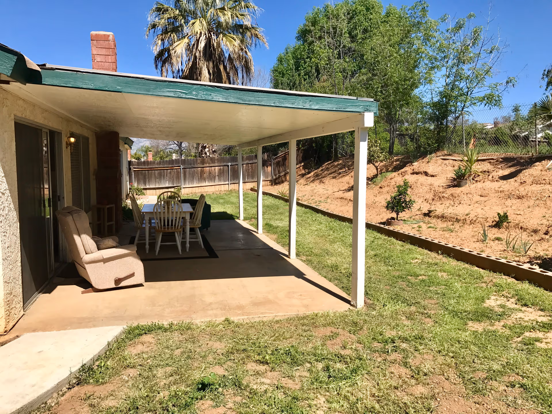 Covered patio with a dining table and recliner overlooking a grassy backyard and sloped planting area.