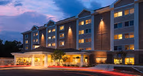 Exterior view of a multi-story assisted living facility building at dusk with lights on inside and outside, featuring a covered entrance and a driveway with light trails from passing vehicles.