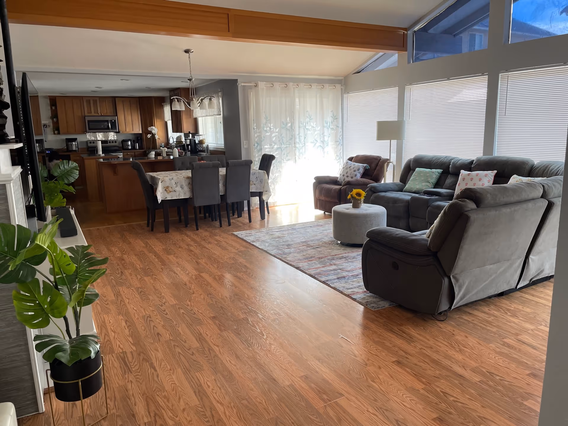 A spacious open living area with wooden flooring featuring a gray sectional sofa, a brown armchair, a round ottoman with a small sunflower vase, and a dining table with six gray chairs covered with a floral tablecloth. The room has large windows with blinds and sheer curtains, allowing natural light to fill the space. The kitchen area with wooden cabinets and stainless steel appliances is visible in the background.