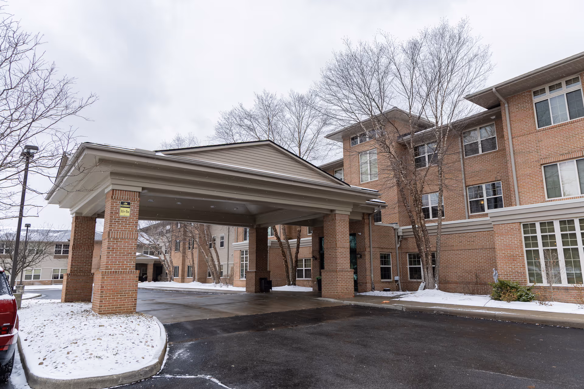 Entrance of a senior living facility with a covered drop-off area supported by brick pillars. The building is made of brick and has multiple windows. There is snow on the ground and leafless trees around the entrance.