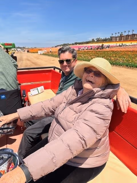An elderly woman wearing a beige sunhat, sunglasses, and a light pink puffer jacket is sitting on a red bench outdoors, smiling at the camera. A man wearing sunglasses and a green jacket is sitting next to her. They appear to be on a vehicle or cart with a dirt path and colorful flower fields in the background under a clear blue sky.