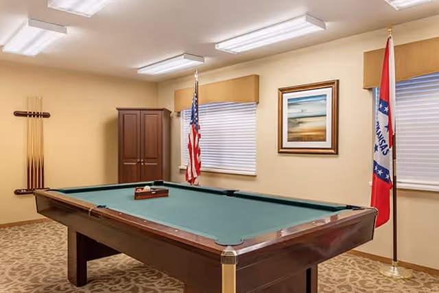 A room with a pool table in the center, featuring a green felt surface and a wooden frame. The room has beige walls and carpeted floor with a patterned design. There are two windows with blinds, an American flag and an Arkansas state flag standing on poles near the windows. A framed landscape picture hangs on the wall between the windows, and a wooden cabinet is positioned in the corner. A rack holding pool cues is mounted on the wall.