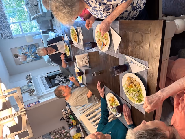 A group of elderly people sitting around a wooden dining table enjoying a meal together in a well-lit room with a fireplace and large window. Plates of food and glasses of dark beverage are on the table, and the room is decorated with paintings and comfortable furniture.