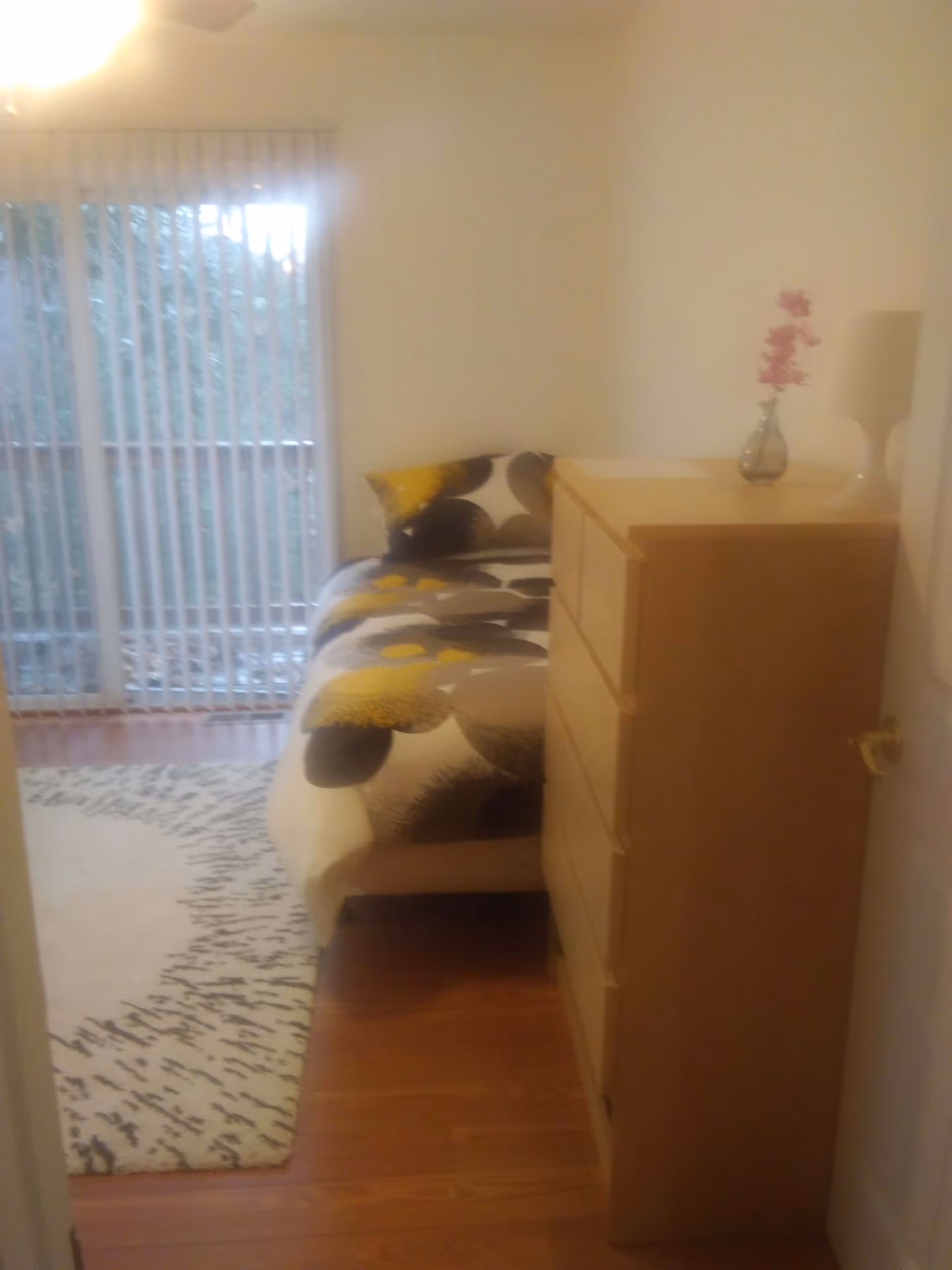 A small bedroom with a bed covered in a patterned duvet featuring yellow, black, and white colors. Next to the bed is a wooden dresser with a small vase holding pink flowers and a white lamp on top. The room has wooden flooring and a large window with vertical blinds letting in natural light.