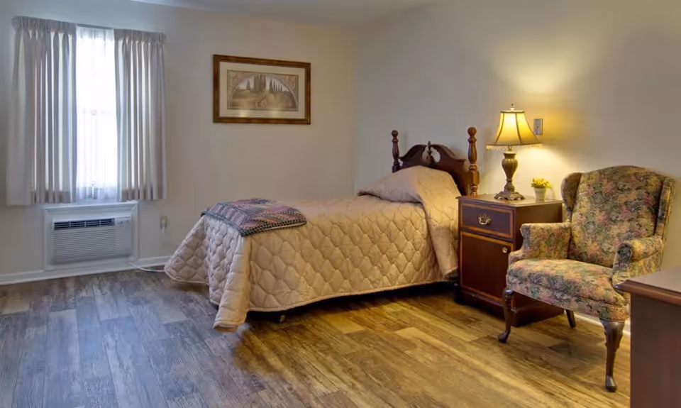 A simple furnished bedroom with a single bed, nightstand and lamp, a floral armchair, framed art on the wall, and a curtained window.