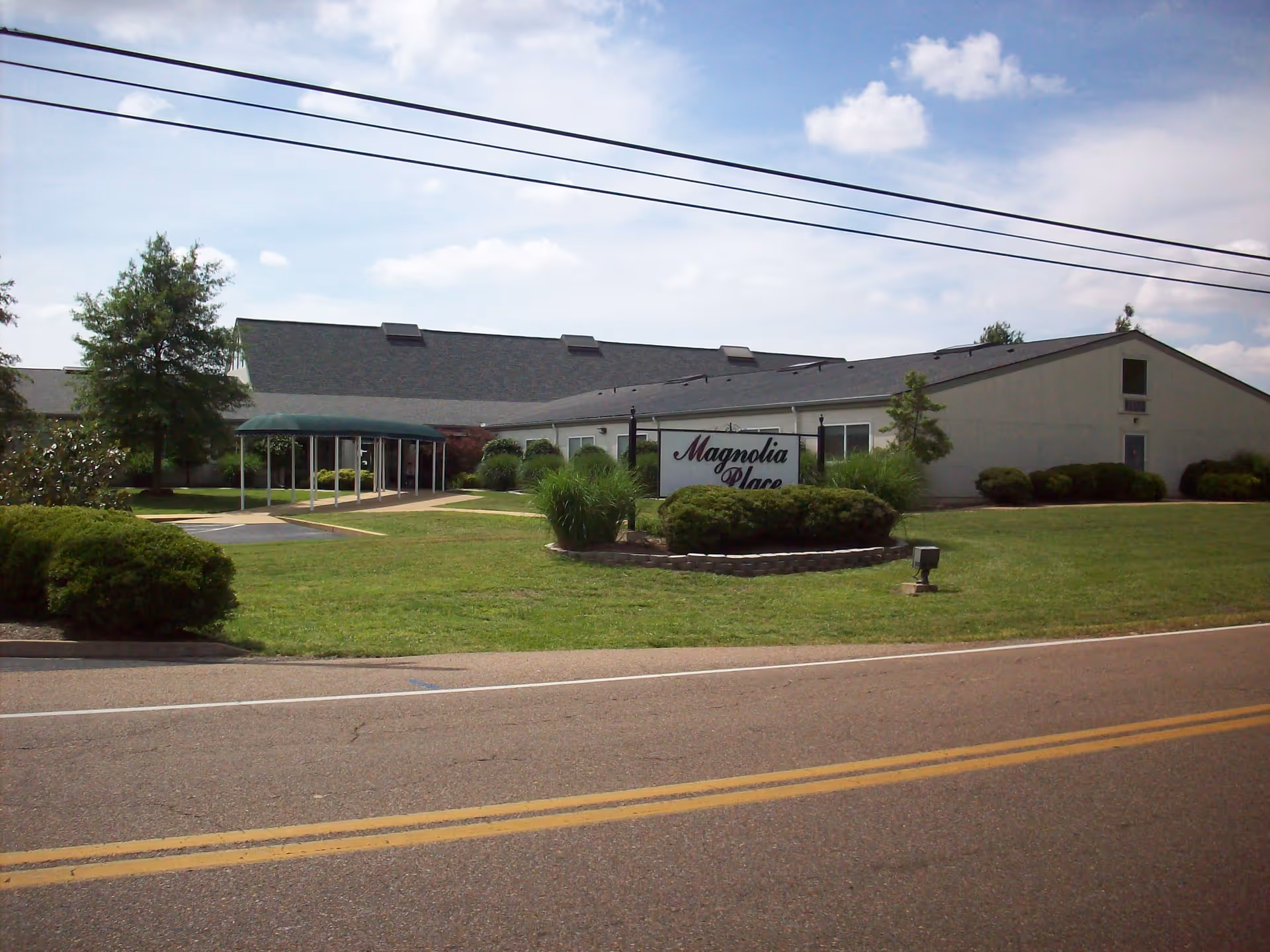 Exterior view of Magnolia Place facility with a single-story building, green lawn, bushes, and a covered walkway leading to the entrance under a partly cloudy sky.