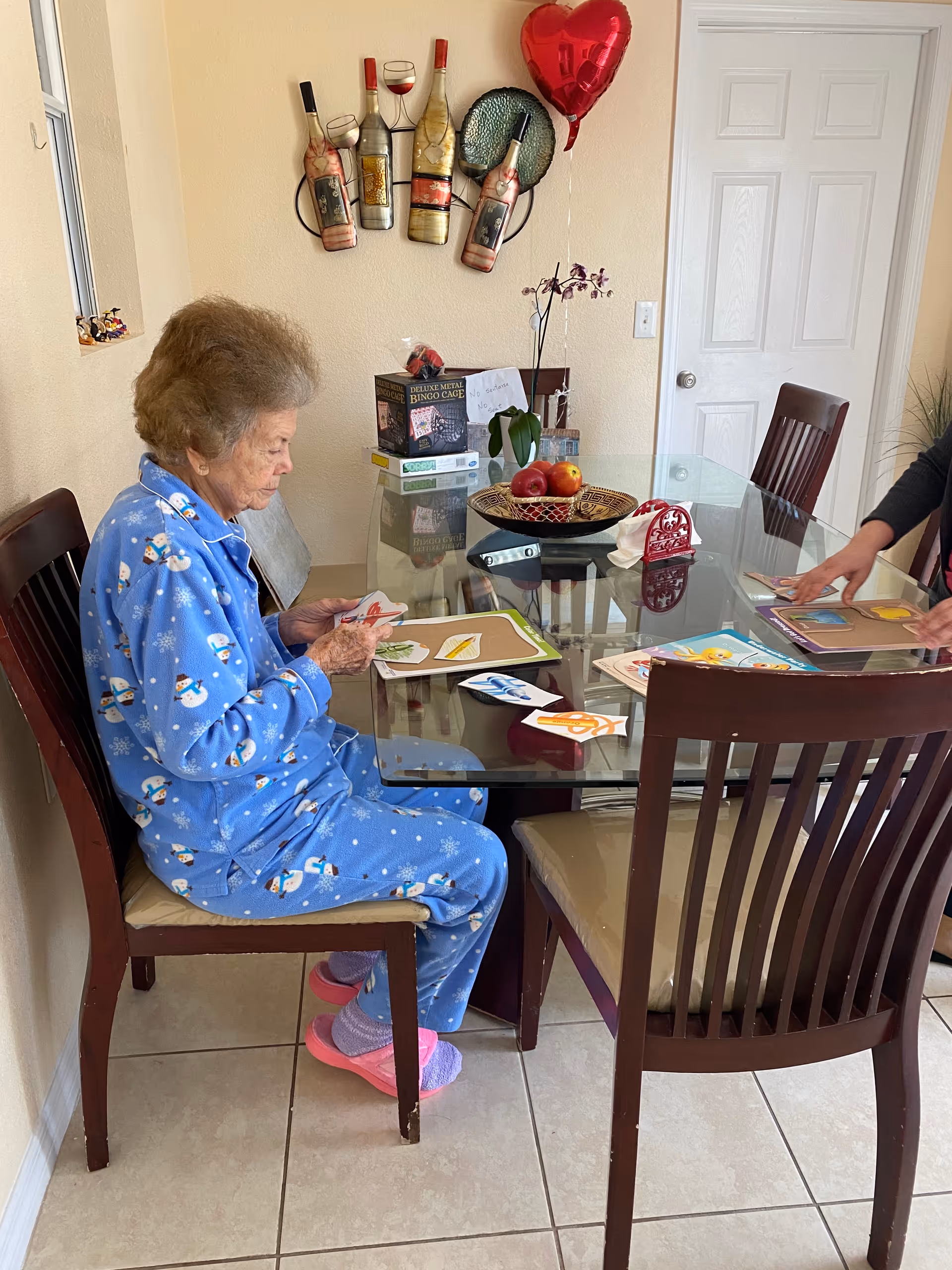 An elderly woman in blue snowman pajamas and pink slippers sits at a glass dining table working on a puzzle or activity with colorful pieces. Another person is partially visible on the right side also working on a similar activity. The room has beige walls, a decorative wine bottle wall hanging, a red heart-shaped balloon, and a bowl of fruit on the table.