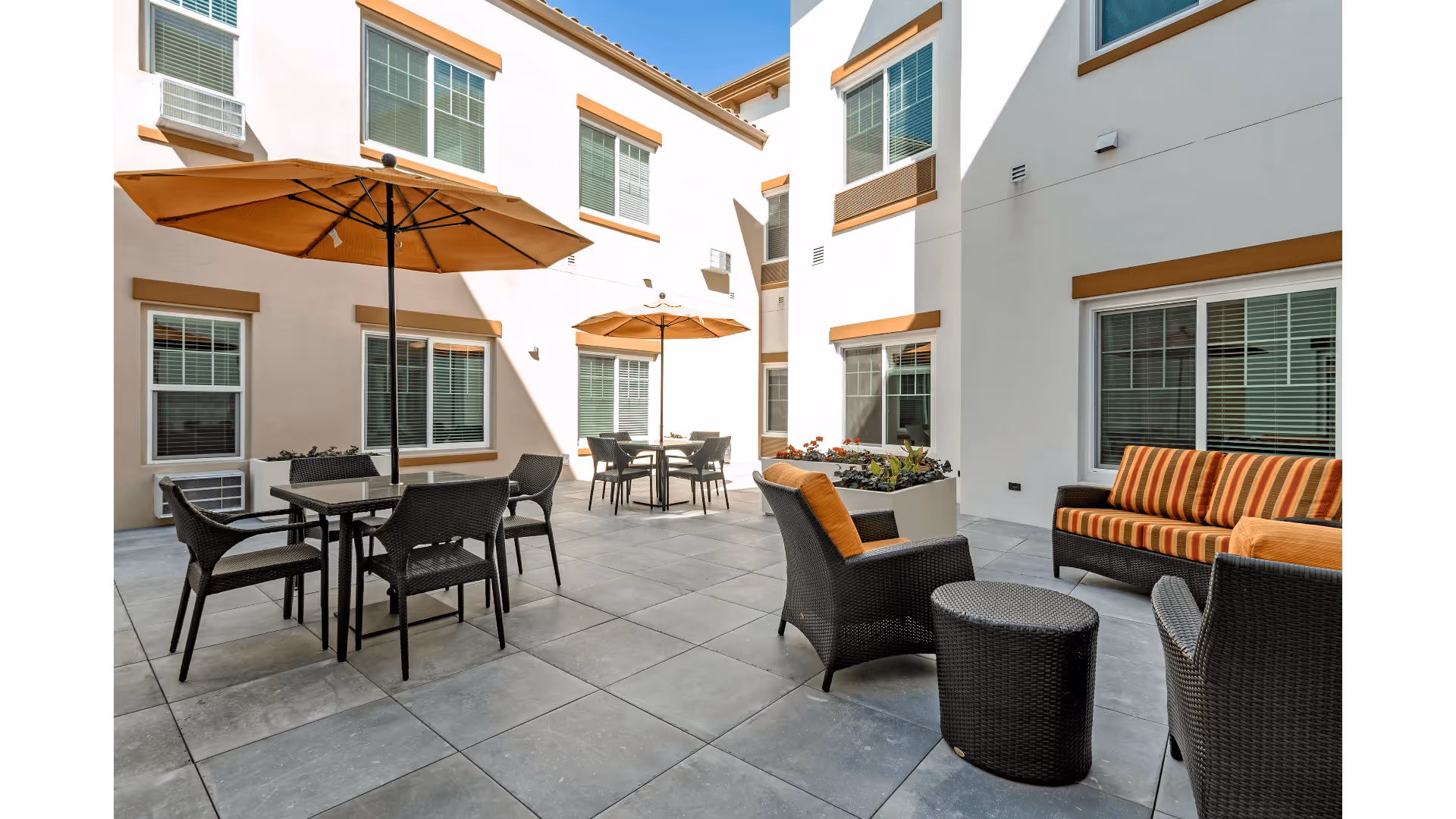 Outdoor courtyard area with patio furniture including tables, chairs, and umbrellas. The space is surrounded by a white building with multiple windows and brown trim. There is a cushioned sofa and chairs with orange and brown striped cushions.