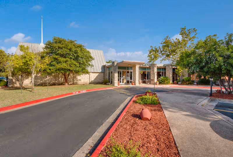 Exterior view of Hilltop Village Nursing and Rehab building with a driveway, landscaped garden beds with red mulch, trees, and a clear blue sky.