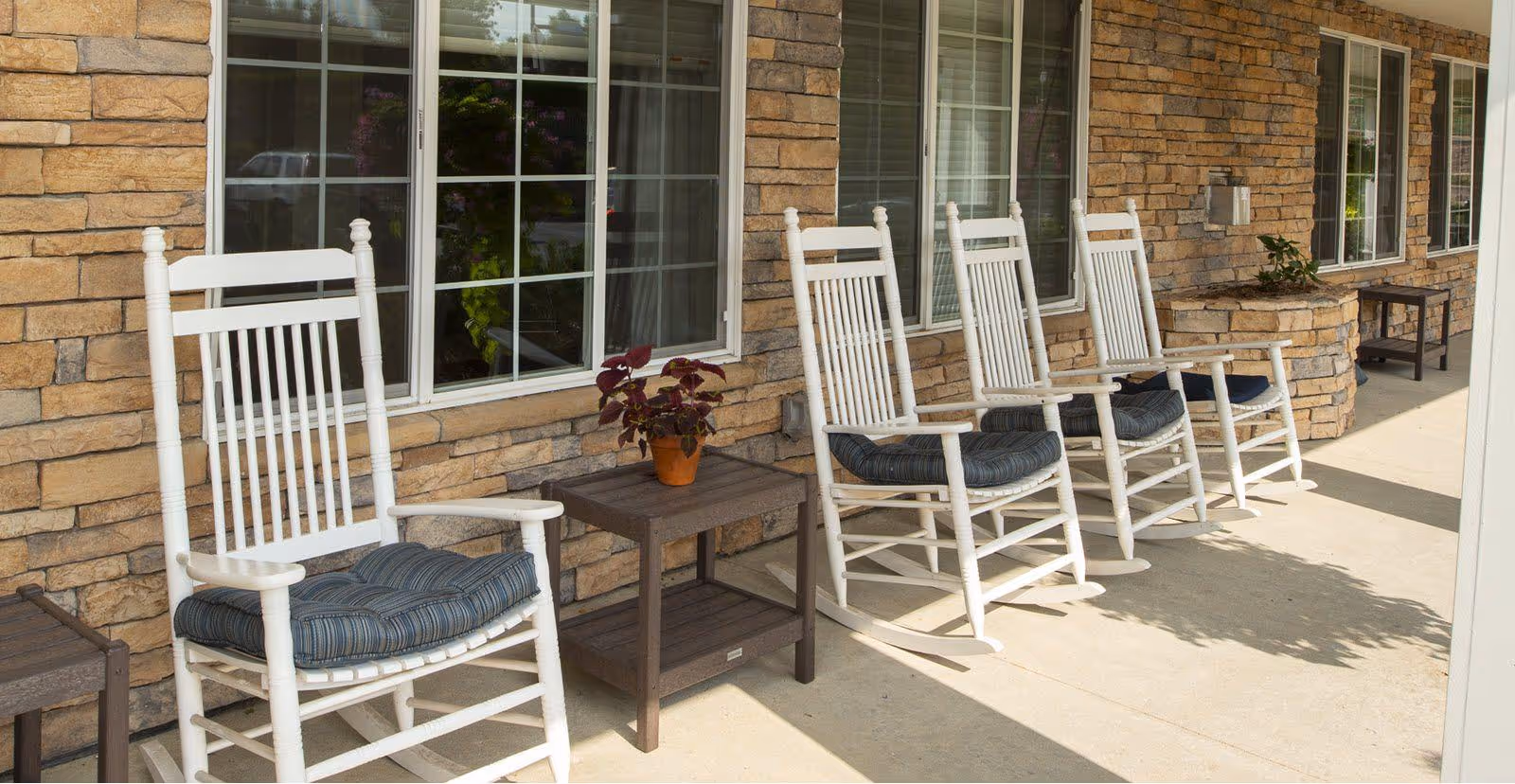 A row of white wooden rocking chairs with blue cushions placed on a concrete patio outside a building with stone walls and large windows. A small wooden side table with a potted plant is positioned between the chairs.