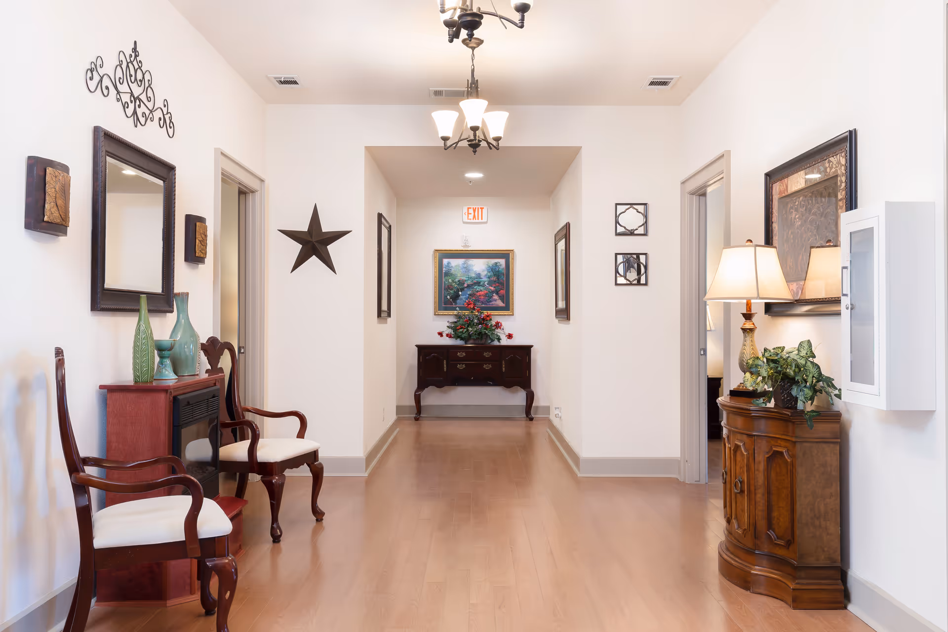 A well-lit hallway in an assisted living facility with wooden flooring and white walls. The hallway features decorative elements including framed artwork, a star wall decoration, a mirror, and wall sconces. There are two wooden chairs with white cushions on the left side, a small red cabinet with green vases, and a wooden side table with a lamp and plant on the right. At the end of the hallway is a dark wooden console table with a floral arrangement and a framed painting above it. An exit sign is visible on the ceiling at the far end.