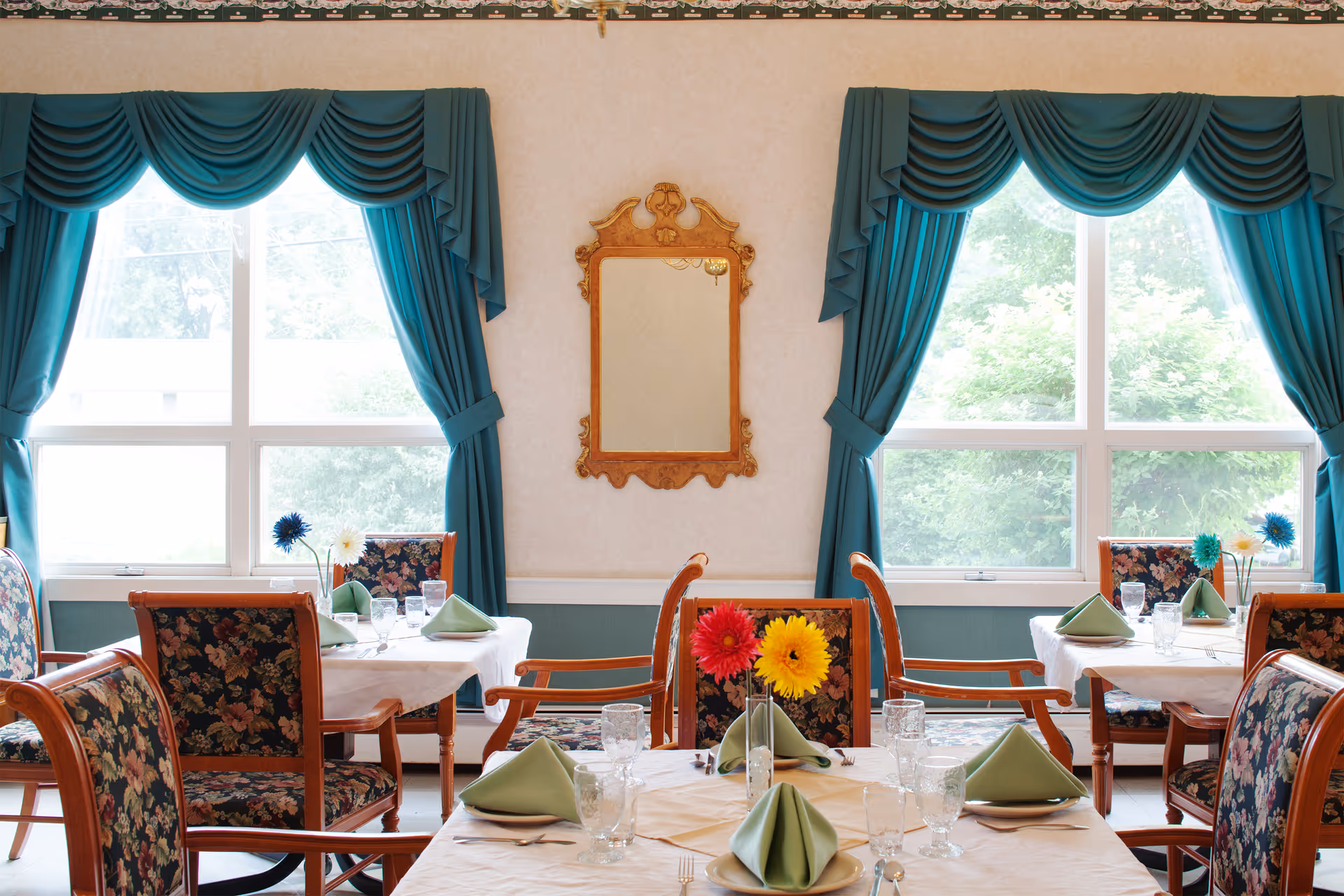 A dining room with tables set for a meal, featuring floral upholstered chairs, white tablecloths, green folded napkins, glassware, and colorful flowers in vases. The room has large windows with teal drapes and a decorative mirror on the wall between the windows.