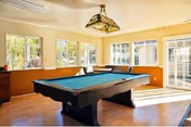 A bright room with large windows letting in natural light, featuring a pool table in the center and a decorative hanging lamp above it. The room has wooden flooring and light-colored walls with a wooden panel halfway up.
