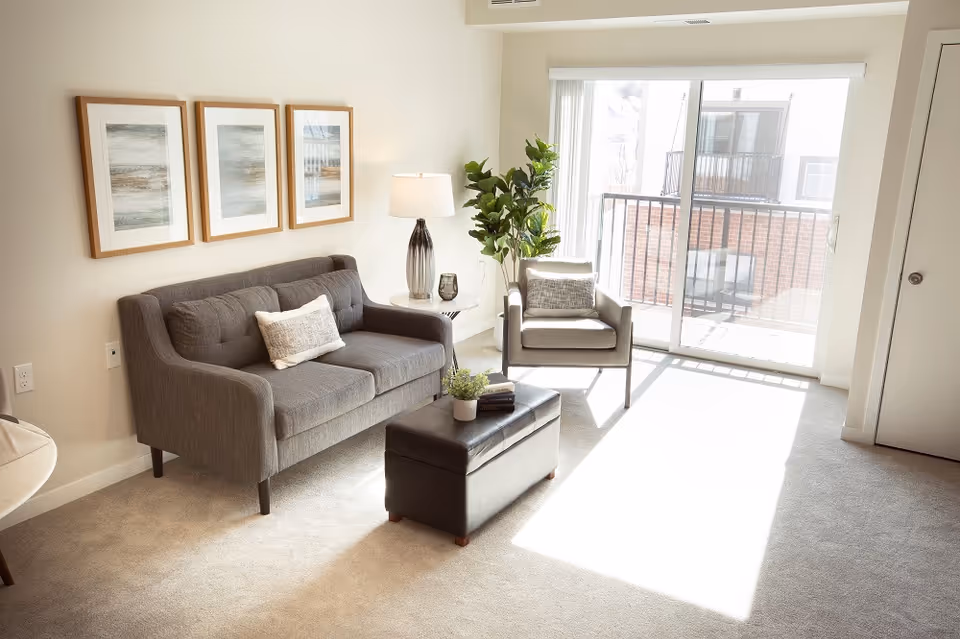 A bright living room with a gray sofa and matching armchair, a small white side table with a lamp, a black ottoman with books and a plant on it, three framed abstract art pieces on the wall, a large potted plant, and a sliding glass door leading to a balcony.