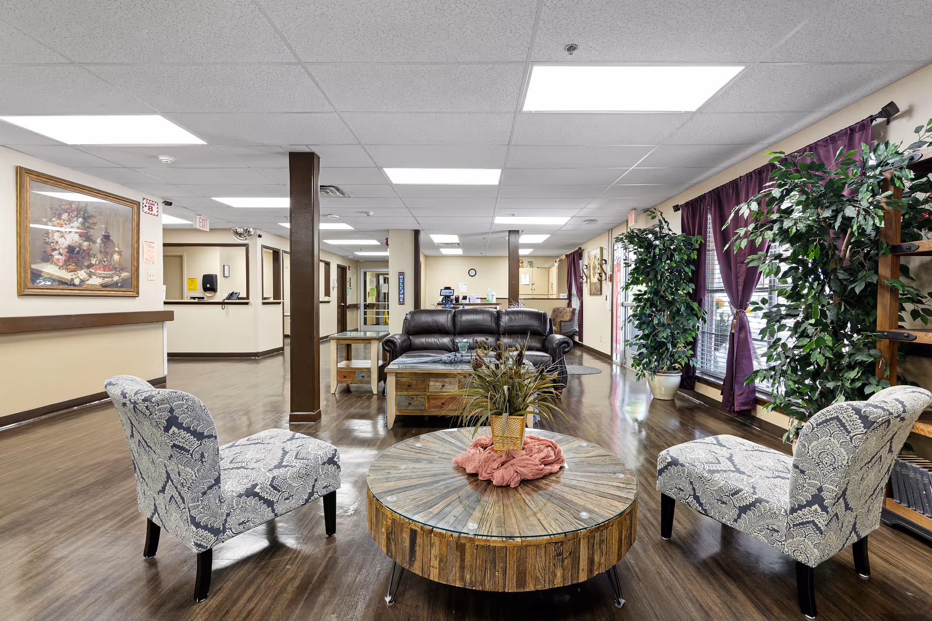 Bright nursing home common lounge with patterned chairs, a round wooden coffee table, a leather sofa, reception desk area, and potted plants by purple-curtained windows.