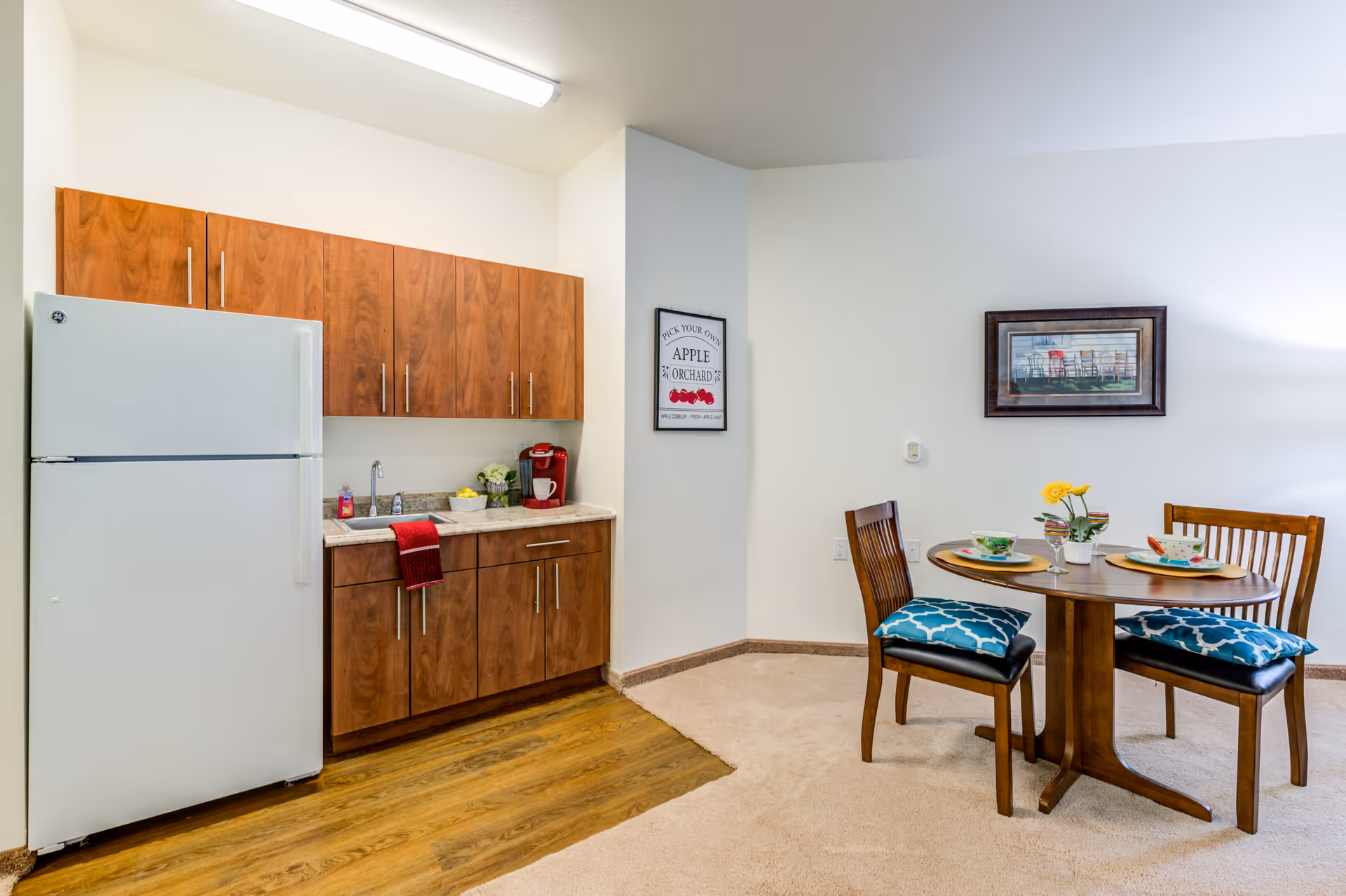 Small kitchenette with wooden cabinets, a white refrigerator and sink next to a round dining table set with two chairs and place settings.