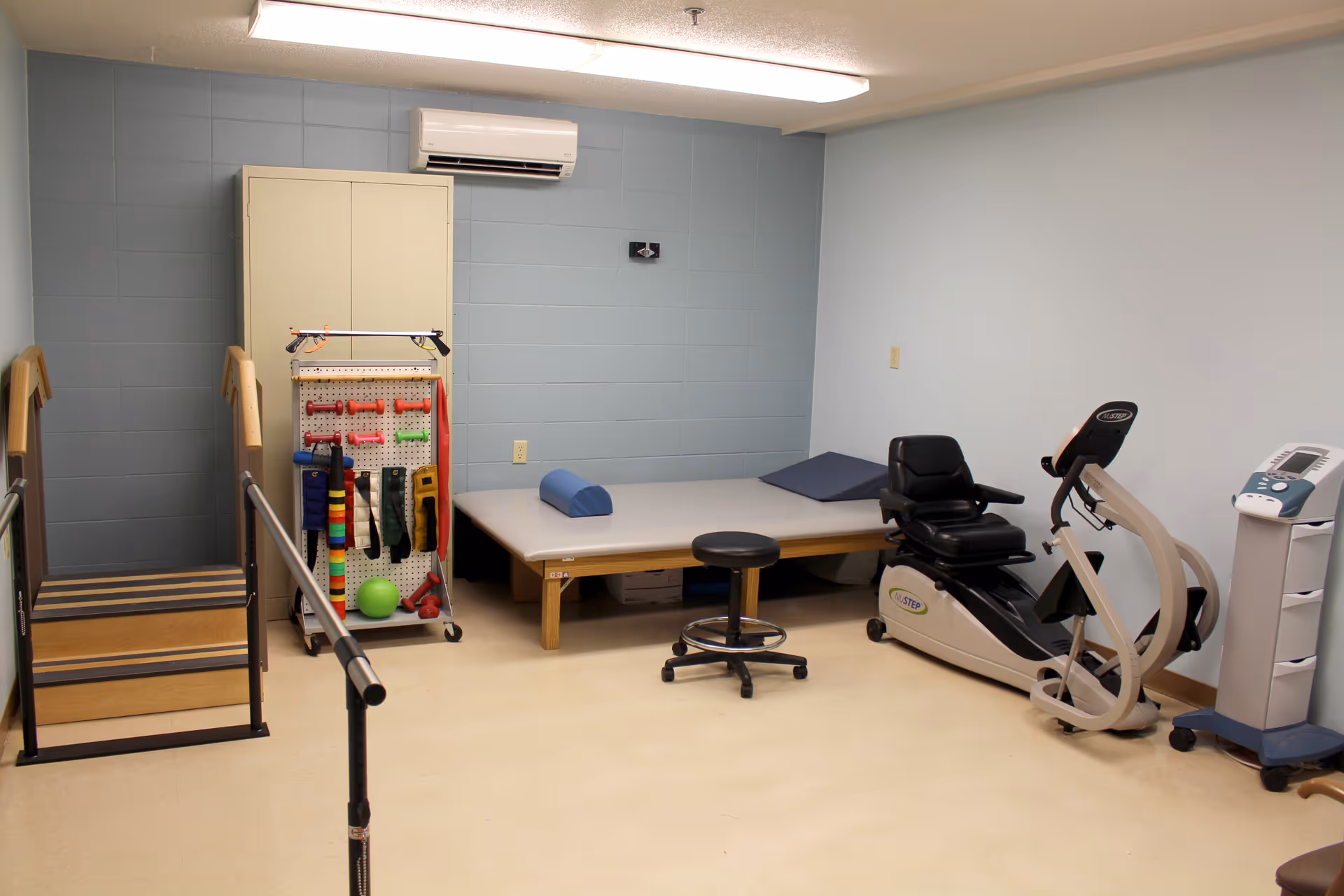 A physical therapy or rehabilitation room with parallel bars for walking exercises, a set of stairs with handrails, a padded therapy table with cushions, various exercise bands and equipment on a pegboard, a black stool, a recumbent exercise bike, and a medical device on a stand. The walls are painted light blue and white, and there is an air conditioning unit mounted on the wall.