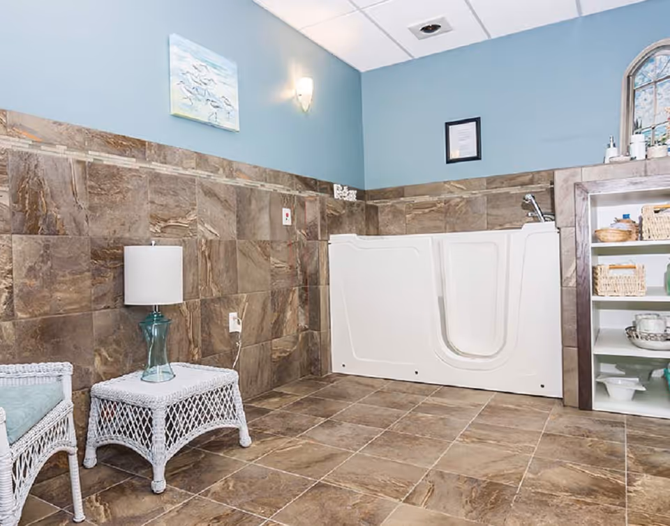 A bathroom with brown tiled walls and floor, featuring a white walk-in bathtub with a door, a wicker side table with a glass lamp, a wicker chair with a cushion, a shelving unit with baskets and toiletries, and light blue painted upper walls with a painting and a framed document.
