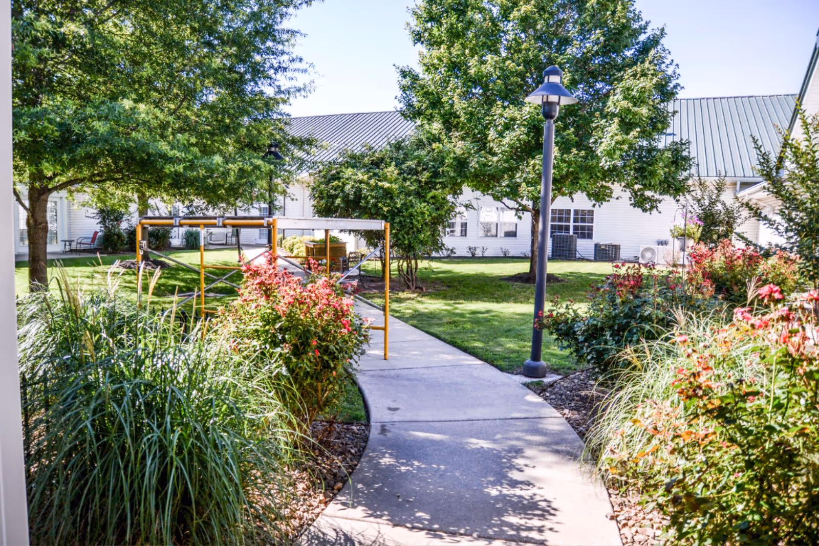 A sunny outdoor garden area with a concrete walkway surrounded by green grass, trees, and flowering bushes. There is a lamppost along the path and white buildings with metal roofs in the background.
