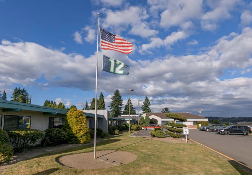 Outdoor view of Mountain View Rehabilitation and Care Center showing a flagpole with the American flag and a flag with the number 12, surrounded by trimmed bushes and a parking lot with several cars under a partly cloudy sky.
