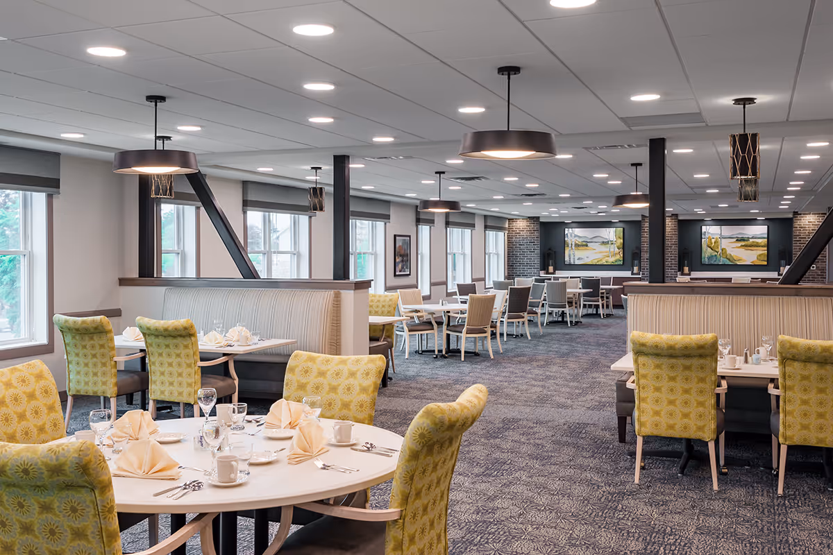 Spacious, well-lit dining room with round and rectangular tables set with napkins and glassware, green patterned chairs, and modern pendant lighting.
