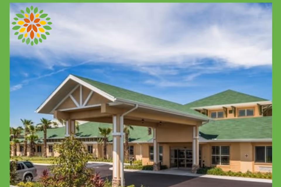 Exterior view of a senior living facility with a covered entrance, beige walls, green roofs, palm trees, and a clear blue sky with some clouds.