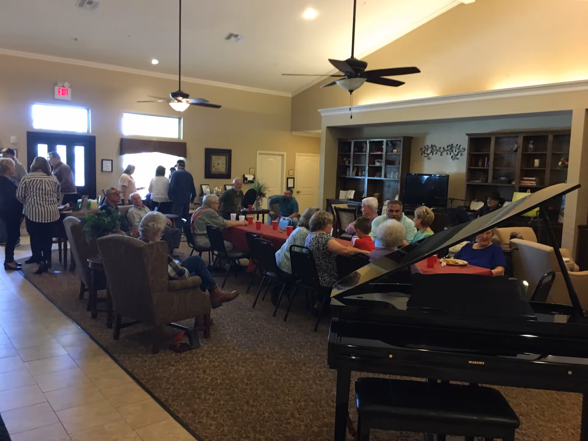 A group of elderly people gathered in a spacious common room with a grand piano in the foreground. Several people are seated around tables covered with red tablecloths, engaging in conversation and enjoying refreshments. The room has ceiling fans, built-in shelves, and a television, creating a comfortable and social atmosphere.
