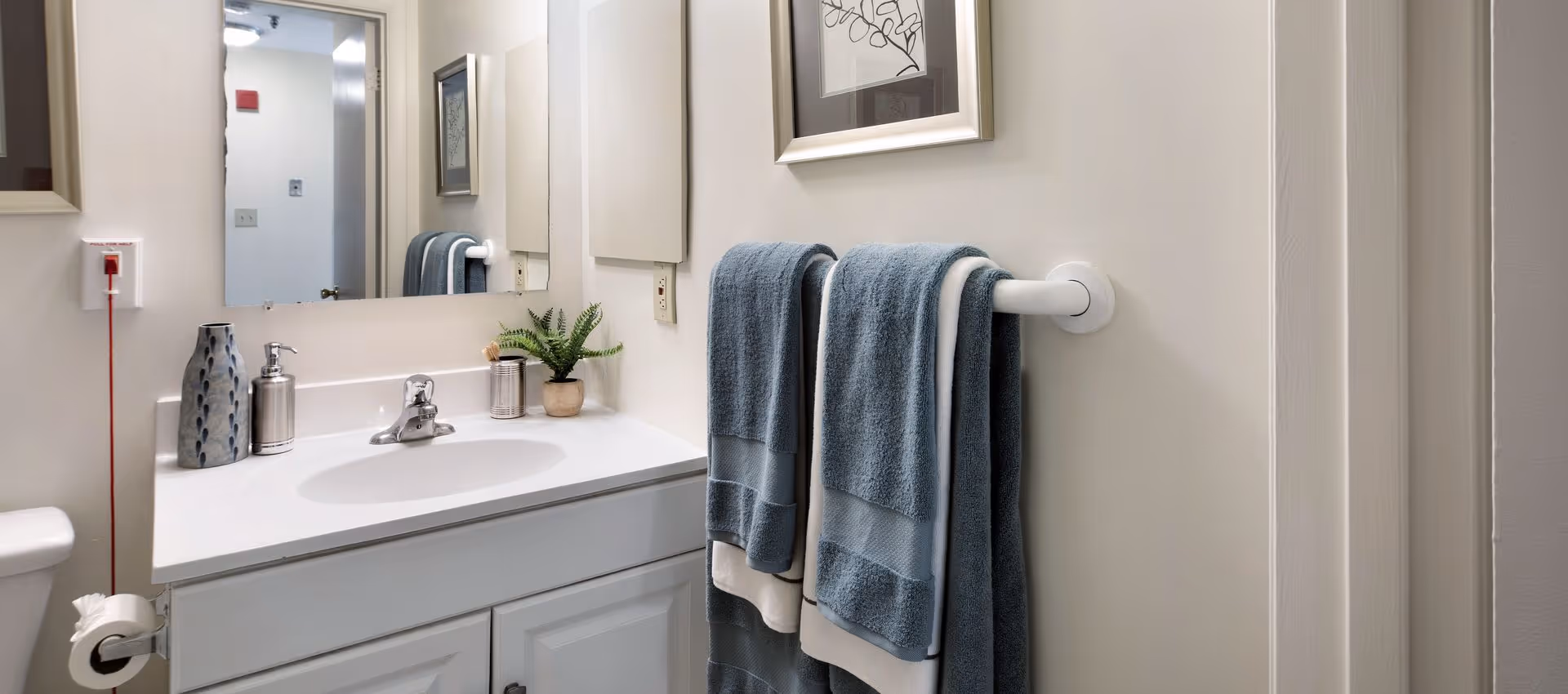 A clean bathroom with a white sink and cabinet, a large mirror above the sink, two blue towels hanging on a white towel rack, a decorative vase, soap dispenser, small plant, and a pull fire alarm on the wall.