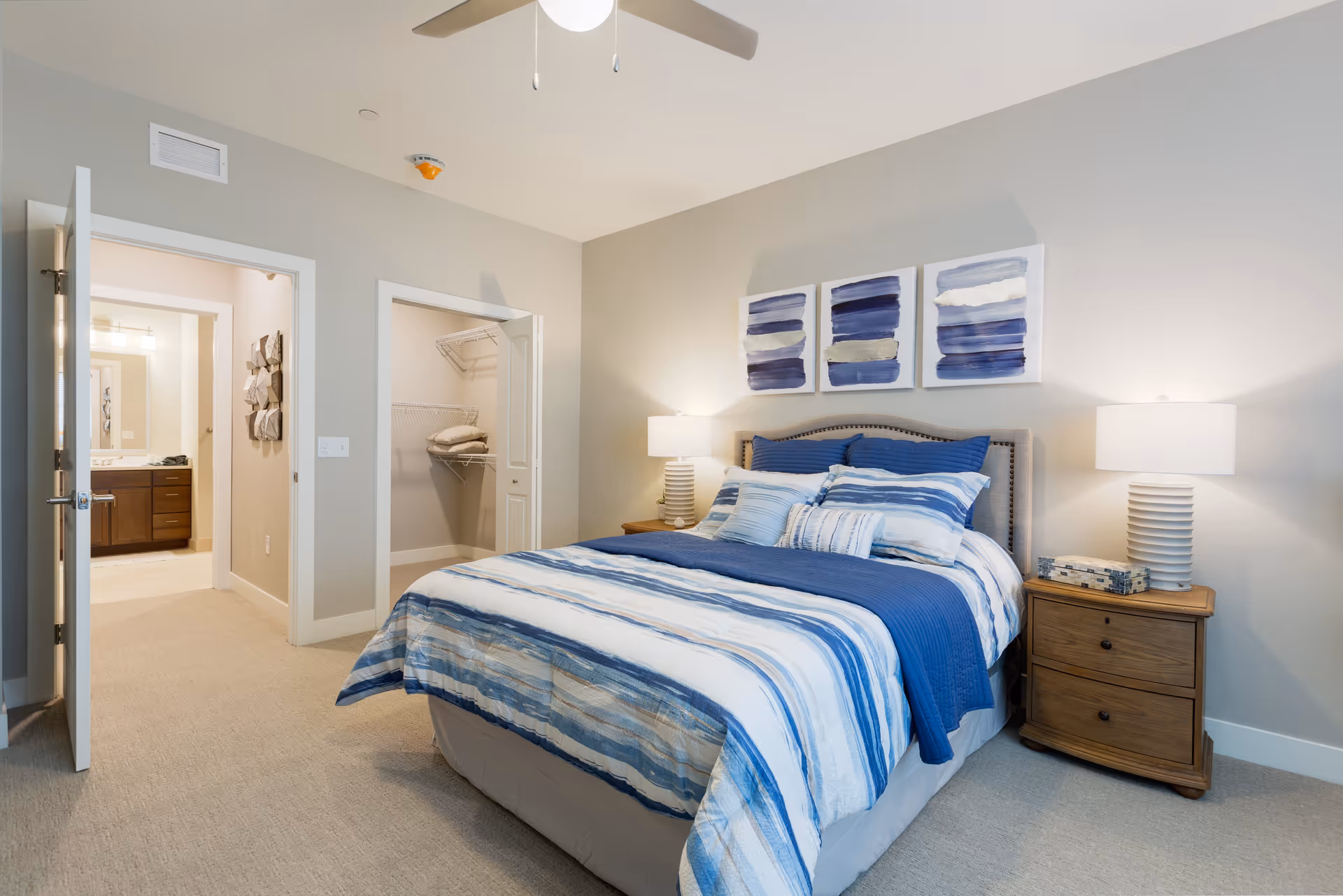 A bedroom with a neatly made bed featuring blue and white striped bedding and multiple pillows. On either side of the bed are wooden nightstands with white lamps. Above the bed are three abstract paintings with blue and white tones. To the left, there is an open door leading to a bathroom and another open door revealing a walk-in closet with shelves and pillows.