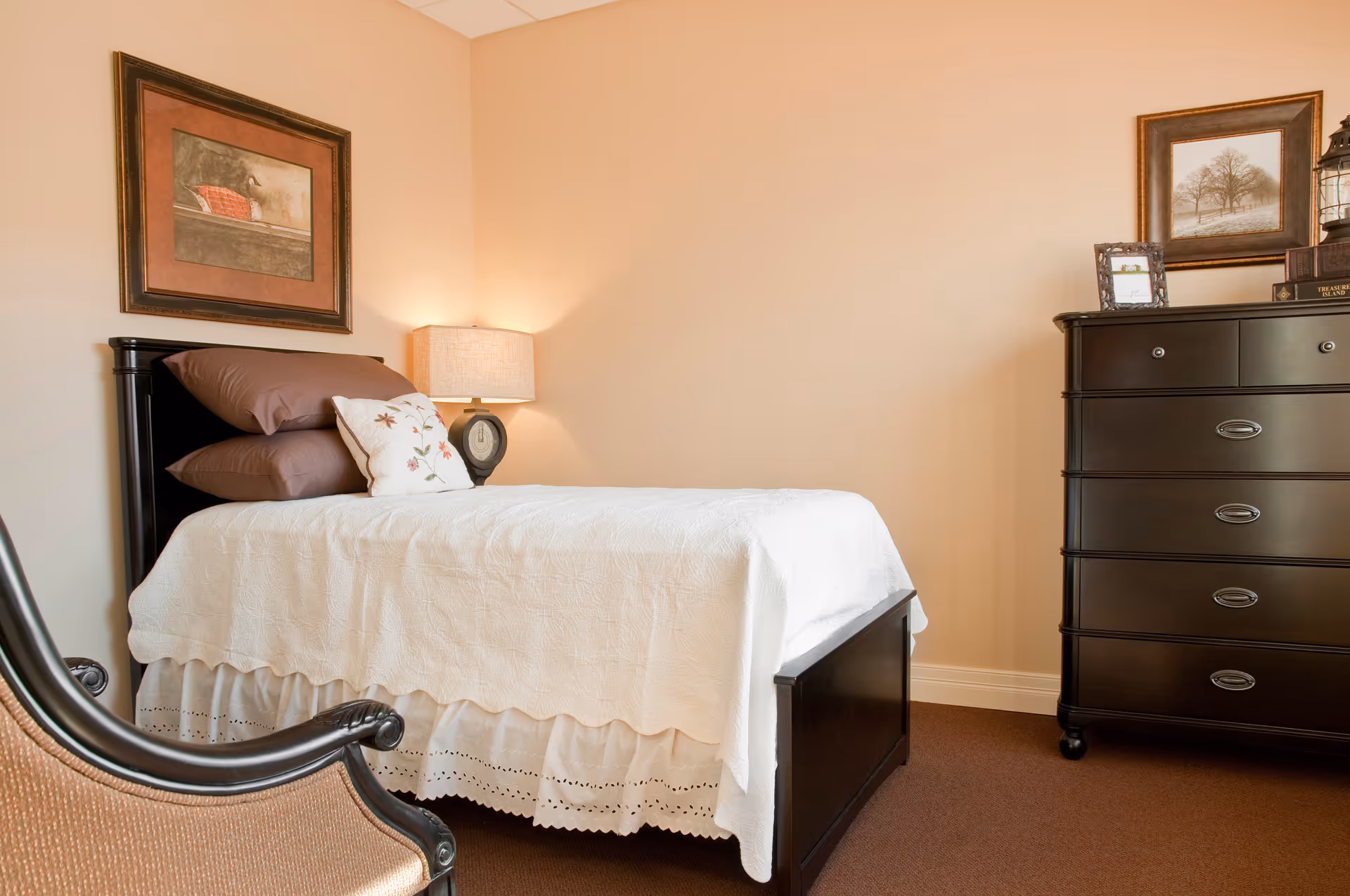 A neatly made single bed with white bedding and brown pillows in a bedroom. There is a bedside table with a lamp and a clock, a dark wooden dresser with framed pictures and decorative items on top, and a framed picture hanging on the wall above the bed. A cushioned chair with wooden armrests is partially visible in the foreground.