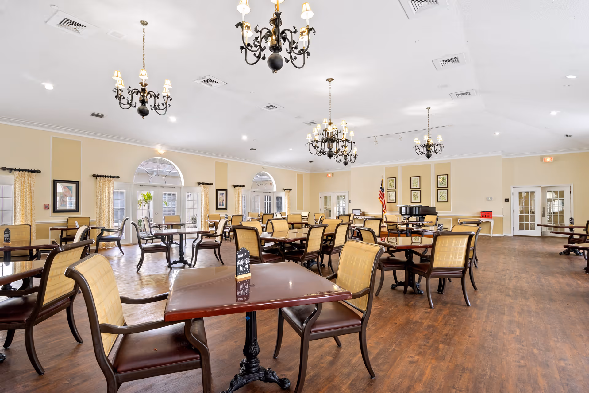 A spacious dining room with multiple wooden tables and chairs arranged neatly on a wooden floor. The room features large arched windows with yellow curtains, several black chandeliers hanging from the white ceiling, framed artwork on the walls, and an American flag near a black grand piano in the corner.