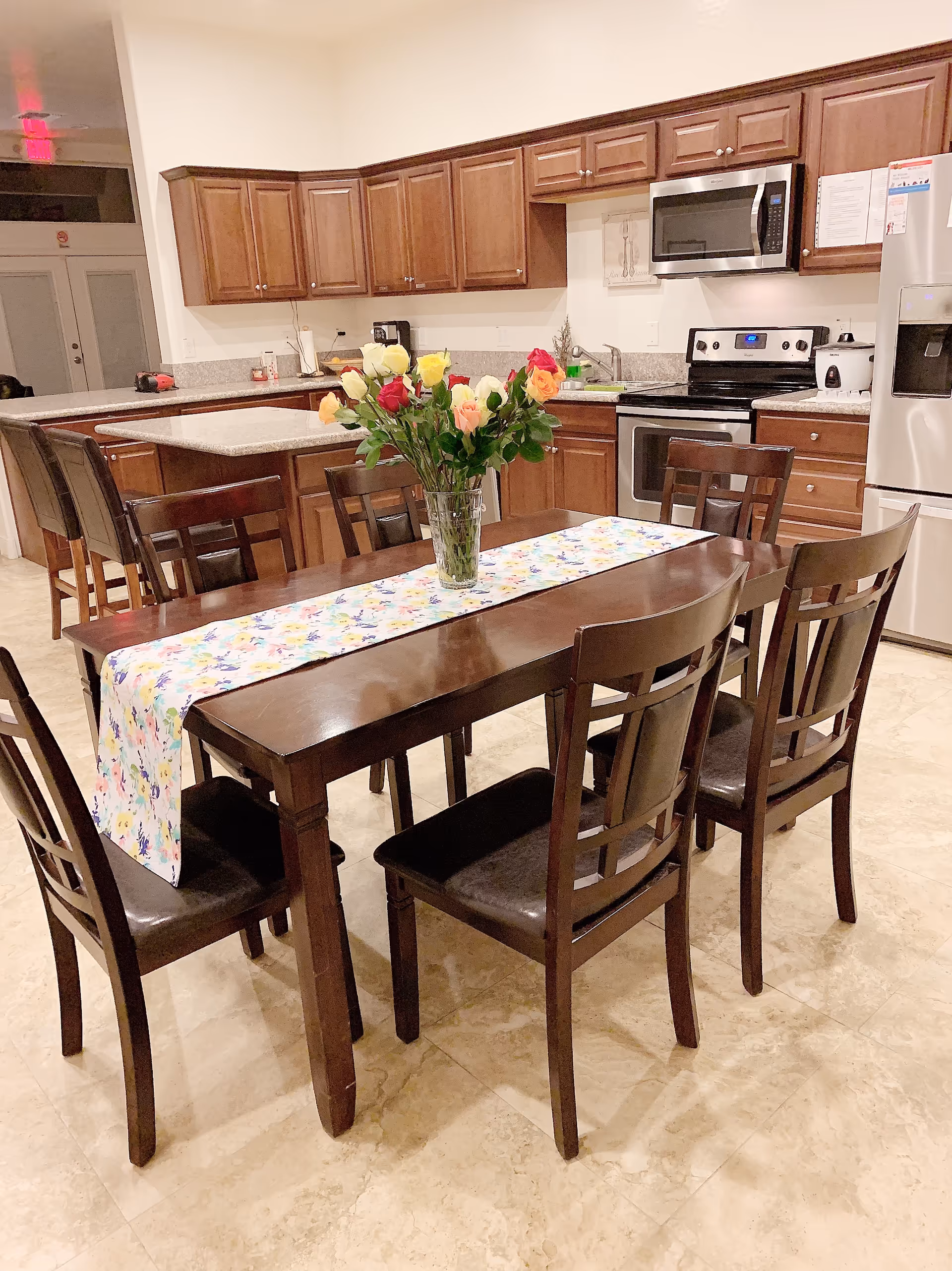 A kitchen and dining area with wooden cabinets, stainless steel appliances including a refrigerator, stove, and microwave. A dark wooden dining table with six matching chairs is in the foreground, decorated with a floral table runner and a vase of colorful roses.