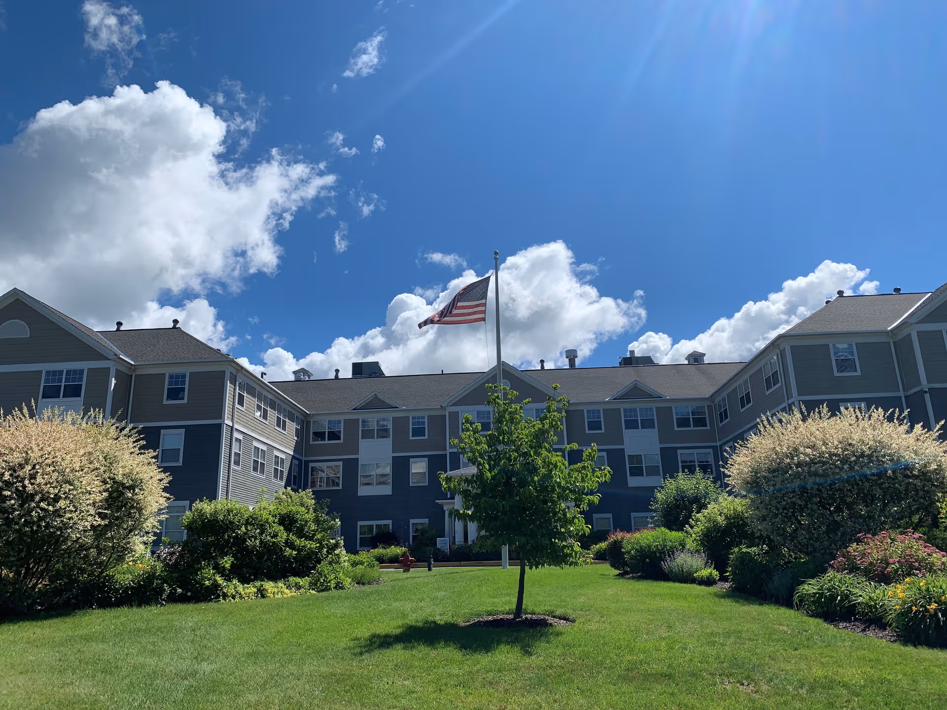 Exterior view of a multi-story senior living facility building with an American flag on a flagpole in the center of a well-maintained green lawn and landscaped bushes under a blue sky with scattered clouds.