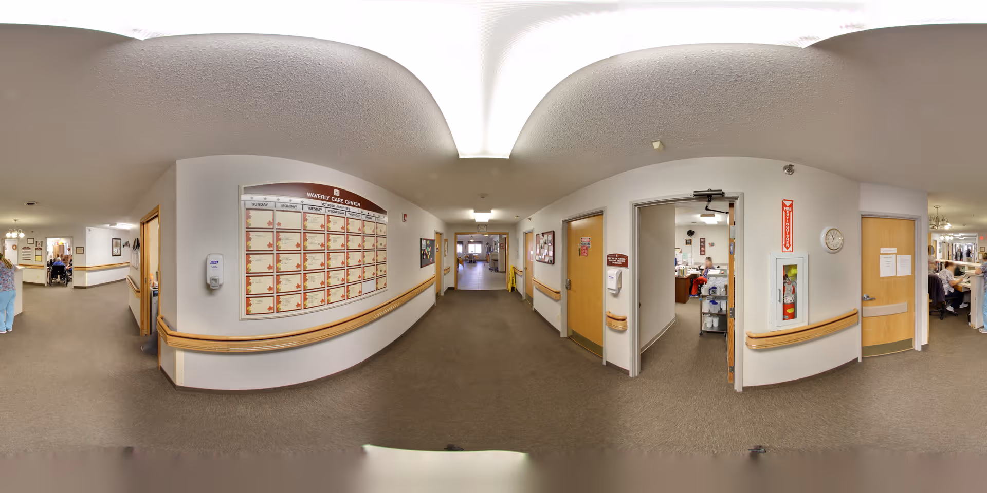 Interior hallway of a senior living facility featuring handrails, a large schedule board, open doors, and people down the corridor.