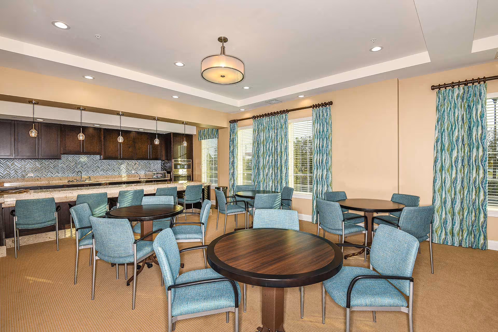 A bright dining room with multiple round wooden tables surrounded by blue upholstered chairs. The room features large windows with patterned blue and green curtains, beige walls, and a ceiling light fixture. In the background, there is a kitchen area with dark wood cabinets, a tiled backsplash, and pendant lights hanging over a counter with bar stools.