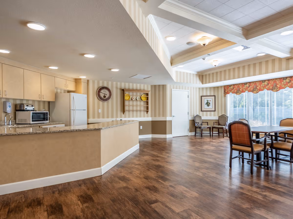 A spacious common area in a senior living facility featuring a kitchen counter with granite countertop, a refrigerator, microwave, and toaster oven. The room has wood flooring, beige and brown striped wallpaper, and recessed lighting. There are several chairs and tables near large windows with floral valances allowing natural light to enter.