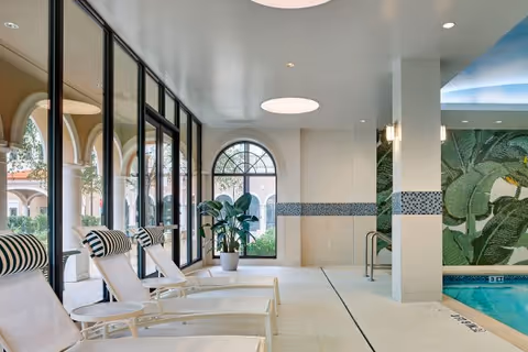 Indoor pool lounge area with white chaise lounges, arched windows, potted plants, and mosaic-tiled walls.