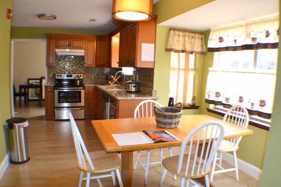 A bright kitchen and dining area with wooden cabinets, stainless steel stove, and granite countertops. The dining table has four white wooden chairs around it, a basket, and an open magazine on top. The room has green walls and windows with coffee-themed curtains letting in natural light.