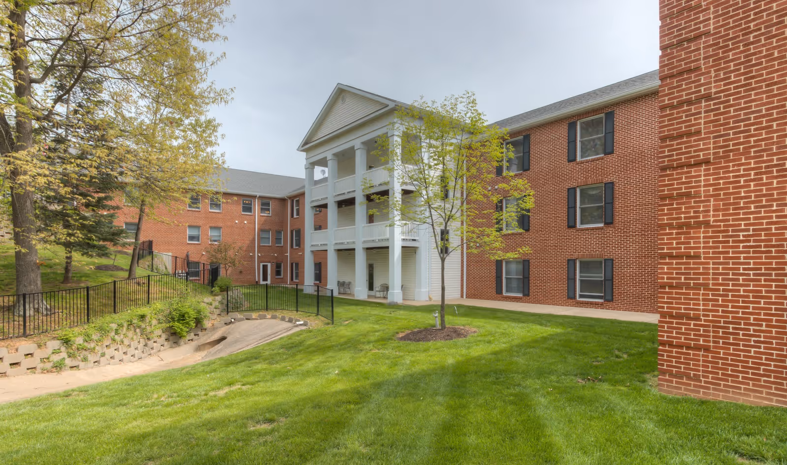 Exterior view of a multi-story brick building with white columns and balconies, surrounded by green grass, trees, and a black metal fence under a cloudy sky.