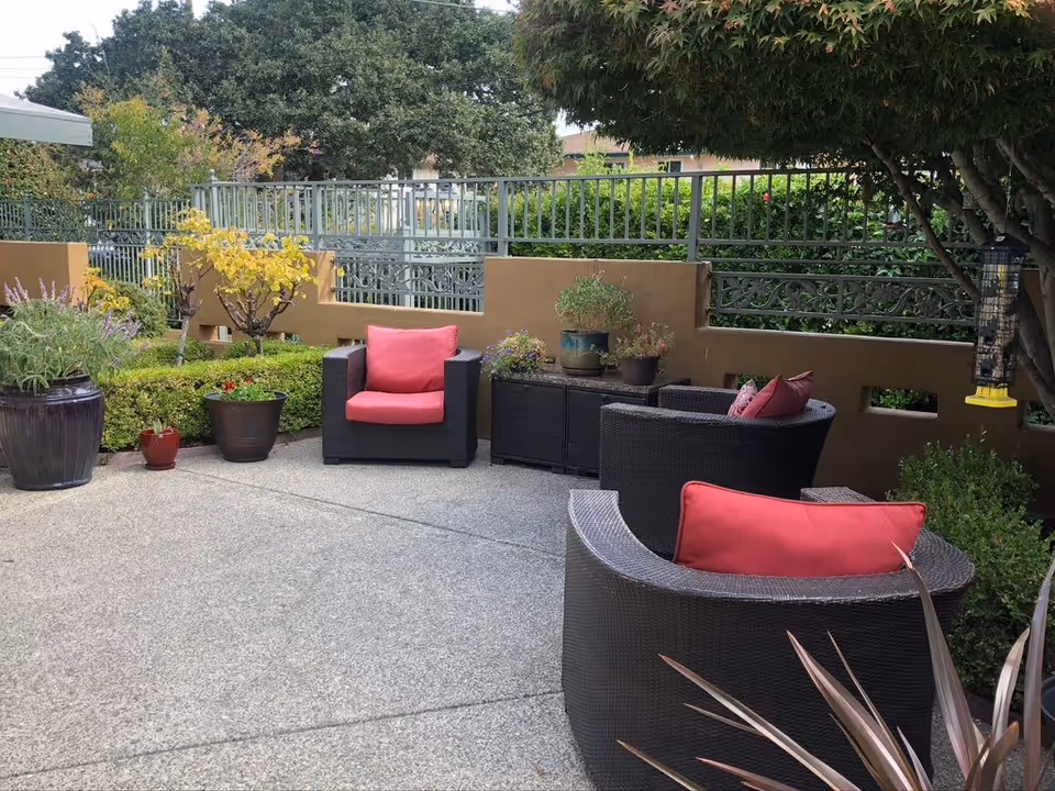 Outdoor patio area with three dark wicker chairs featuring red cushions arranged around a small black table. The patio is surrounded by various potted plants and greenery, with a metal fence and trees in the background.