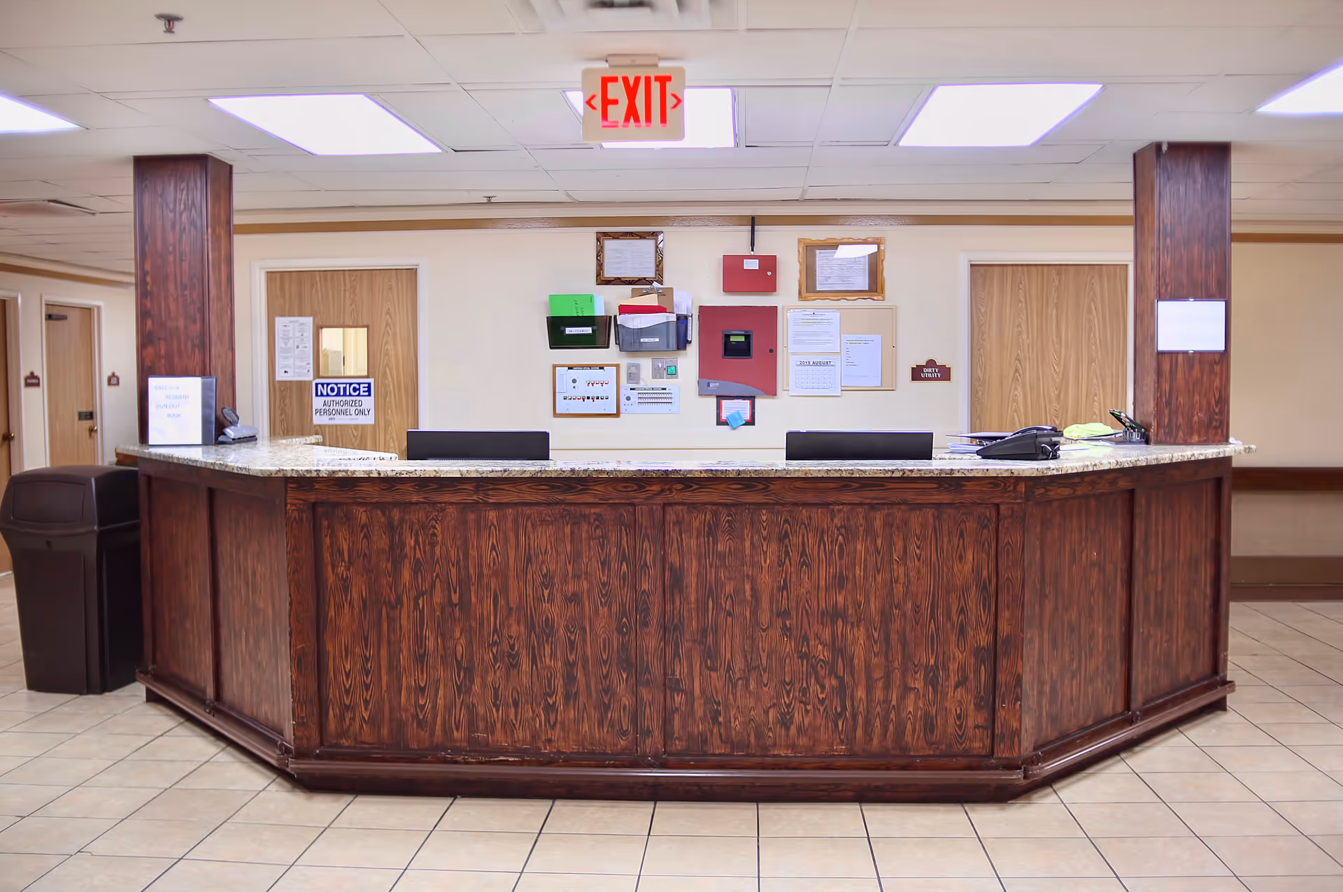 Reception desk area with a wooden counter and granite top in a nursing center. Behind the desk are two wooden doors, various notices and documents pinned on the wall, and an illuminated red exit sign above. The floor is tiled and the ceiling has fluorescent lights.