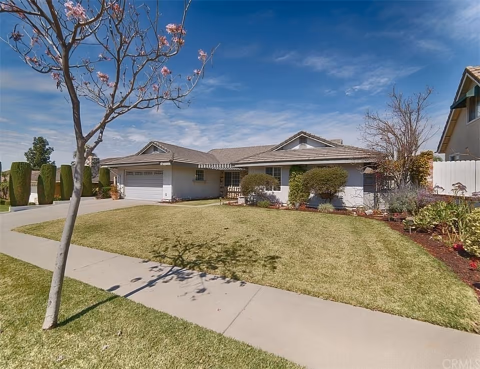 Single-story suburban house with a front lawn, driveway and a blossoming tree under a blue sky.