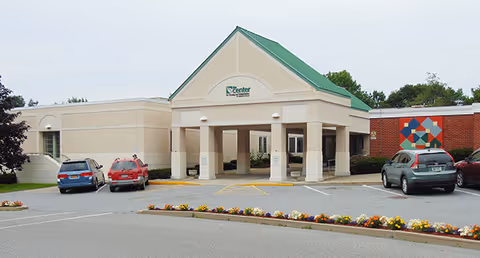 Exterior view of The Center For Nursing And Rehab building with a covered entrance, several parked cars, and a flower bed in the foreground.