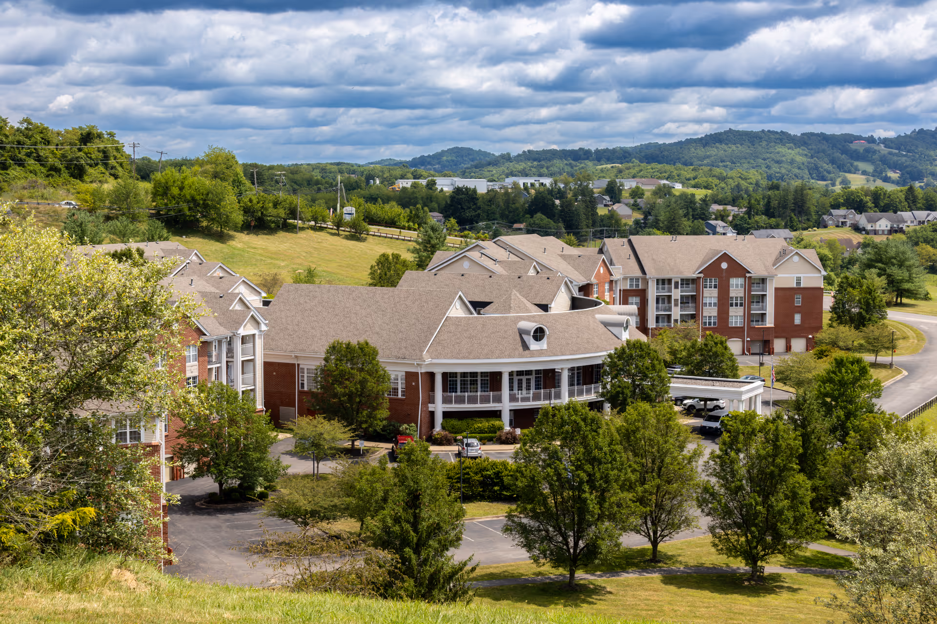 Aerial view of a senior living facility complex with multiple connected buildings surrounded by green trees and grassy areas, set against a backdrop of rolling hills and a partly cloudy sky.