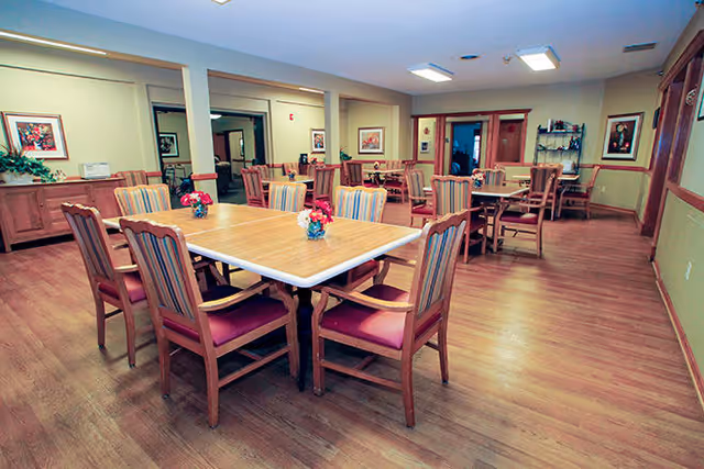 A spacious dining room with multiple wooden tables and chairs arranged neatly. Each table has a small floral centerpiece. The room has wooden flooring, beige walls with framed artwork, and ceiling lights providing bright illumination.